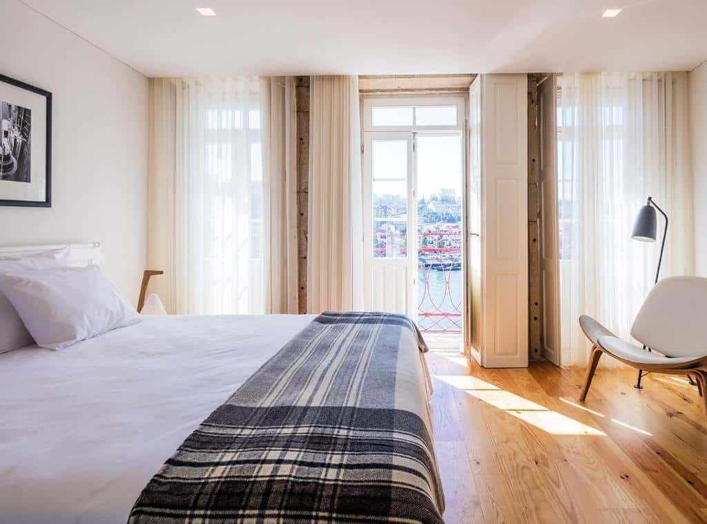 Quarto do Porto River com cama de casal do lado esquerdo da imagem, em frente a cama do lado esquerdo da cama uma cadeira e atrás da cadeira portas que dá acesso a varanda com vista para o Douro. Representa hotéis com vista do Douro.