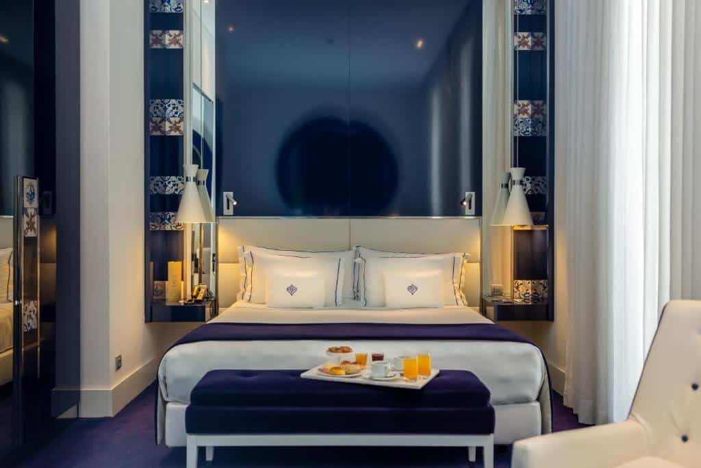 Quarto do Portugal Boutique Hotel com uma cama de casal, uma janela com cortinas, uma poltrona branca, um banco ao pé da cama com uma bandeja com café da manhã, para representar hotéis no Chiado em Lisboa