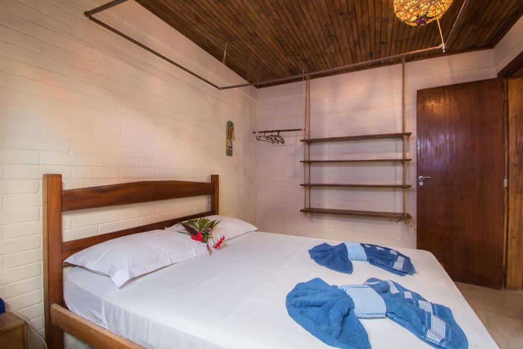 Quarto da Pousada Azul. Uma cama de casal na frente, atrás um lugar para pendurar roupas com cabide e do lado várias prateleiras. No lado direito, no fundo, a porta do quarto. Foto para ilustrar post sobre Pousadas em Cumuruxatiba (BA).
