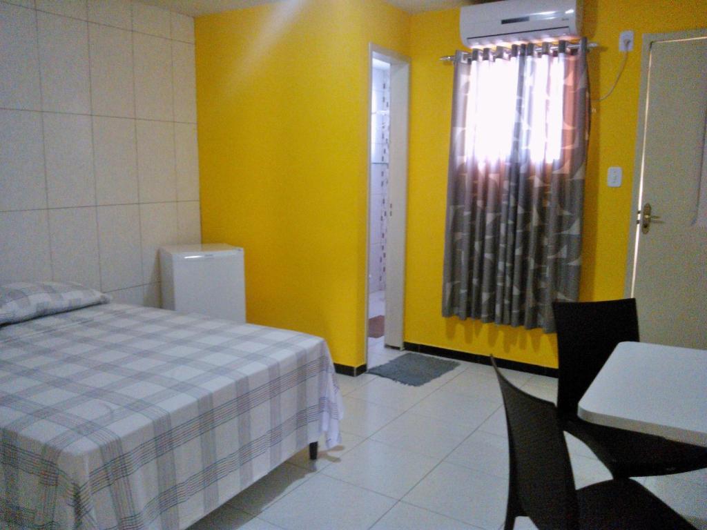 Quarto da Pousada Casa Bella. Uma cama de casal e um frigobar do lado esquerdo. Do lado direito, uma mesa com duas cadeiras. Atrás no lado direito a porta do quarto e a janela. No lado esquerdo, no fundo, a porta do banheiro.