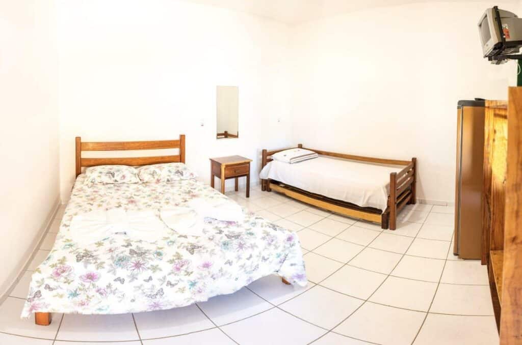 Quarto da Pousada Casarão. Uma cama de casal no lado esquerdo, uma cômoda no meio com um espelho. No lado direito uma cama de solteiro. De frente ara a cama uma geladeira, um armário e uma televisão. Foto para ilustrar post sobre Pousadas em Cumuruxatiba (BA).