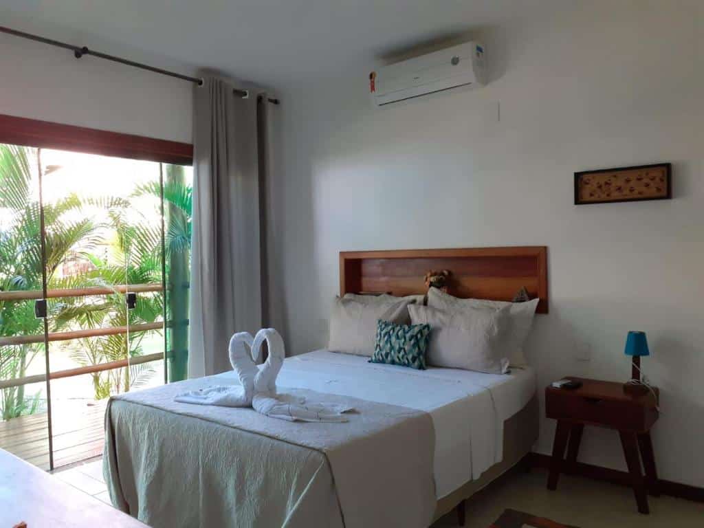 Quarto da Pousada Maré. Uma cama de casal com uma decoração de coração com toalhas. No lado direito da cama um criado mudo com um abajur. No lado esquerdo uma porta de vidro para a sacada. Foto para ilustrar post sobre Pousadas em Cumuruxatiba (BA).