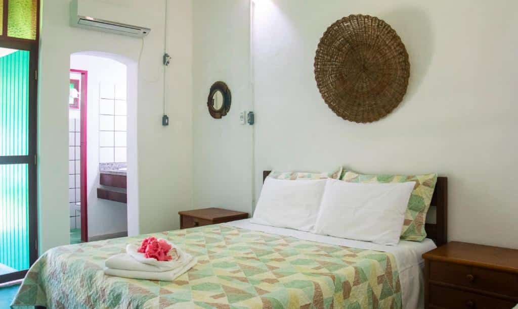 Quarto da Pousada São Francisco. Uma cama de casal no lado direito, com uma cômoda de cada lado. Atrás, para o lado esquerdo, a porta do quarto e a porta do banheiro. Foto para ilustrar post sobre Pousadas em Cumuruxatiba (BA).