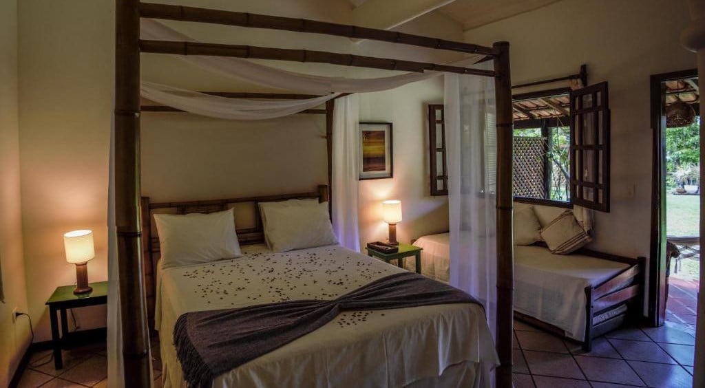 Quarto da Pousada Villa Cumuru. Uma cama de casal com dossel. De cada lado da cama um abajur. No lado direito a porta do quarto e em baixo da janela um sofá cama. Foto para ilustrar post sobre Pousadas em Cumuruxatiba (BA).