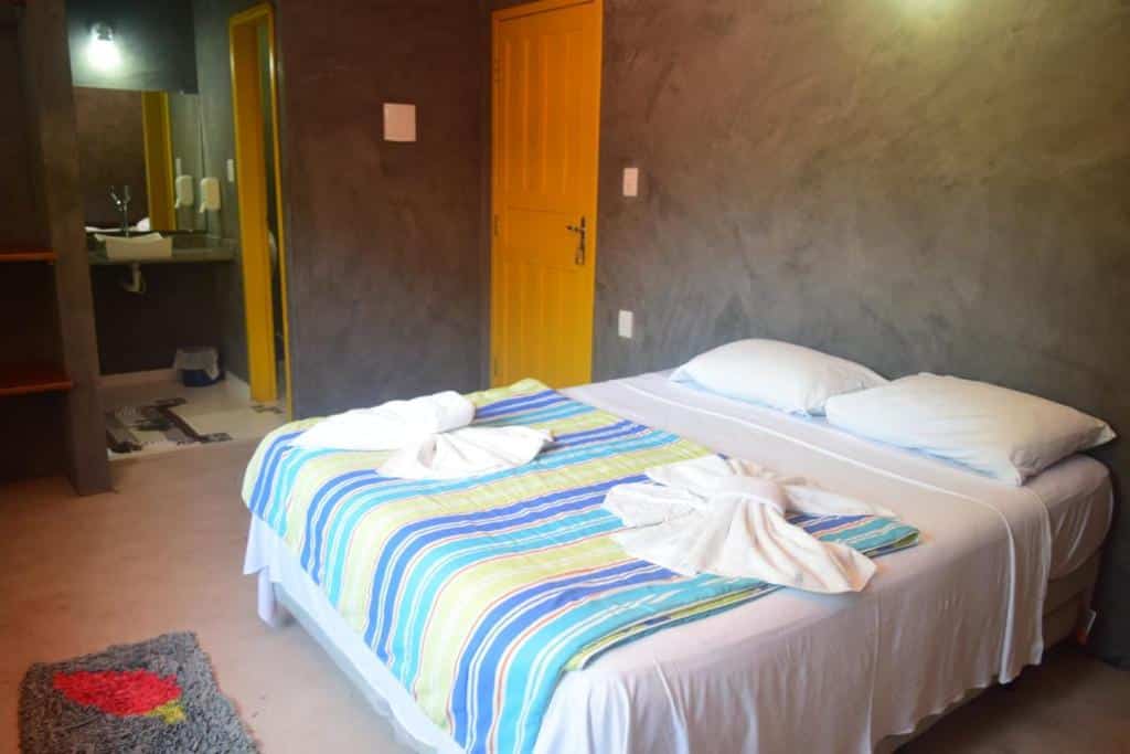 Quarto da Pousada Zen. Do lado direito uma cama de casal na frente, com duas toalhas decorando a cama. Do lado esquerdo da cama a porta do quarto. No lado esquerdo do quarto a porta do banheiro com uma pia e um espelho.