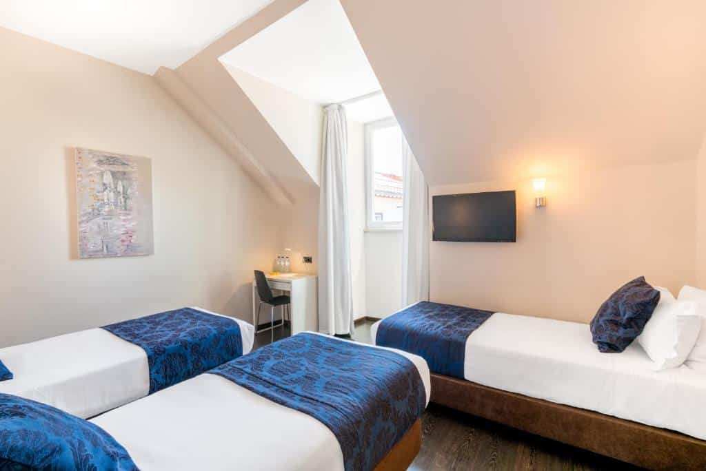 Quarto do Rossio Garden Hotel com três camas de solteiro, uma janela com cortinas, uma mesinha de escritório com uma cadeira e uma televisão, para representar hotéis para família em Lisboa