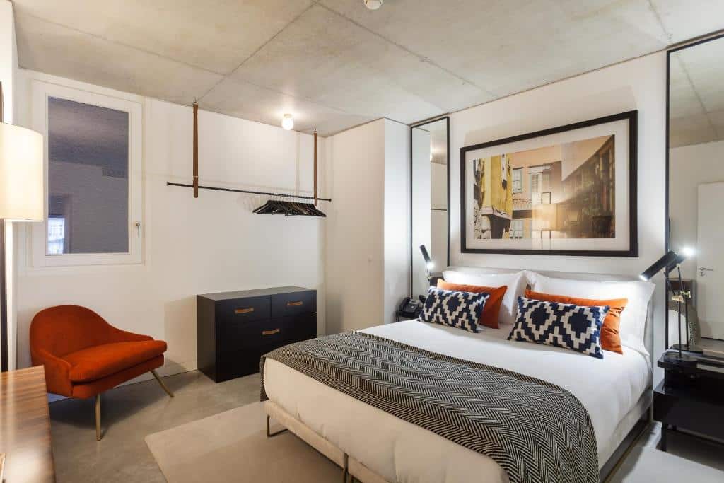 Quarto do S.Bento Residencescom cama de casal do lado direito da imagem, do lado esquerdo da cama uma cômoda escura e uma poltrona laranja.