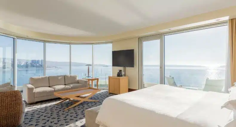 Quarto no Sheraton Miramar Hotel & Convention Center. Há uma cama de casal posicionada no meio, um sofá do outro lado e virado para ela. Entre eles há um recamier ao pé da cama e uma mesa de centro. Do lado esquerdo do sofá há uma poltrona e do lado direito um pequeno armário e tv. O quarto é quase todo com paredes de vidro e vista para o mar. Este é um dos hotéis em Viña Del Mar.