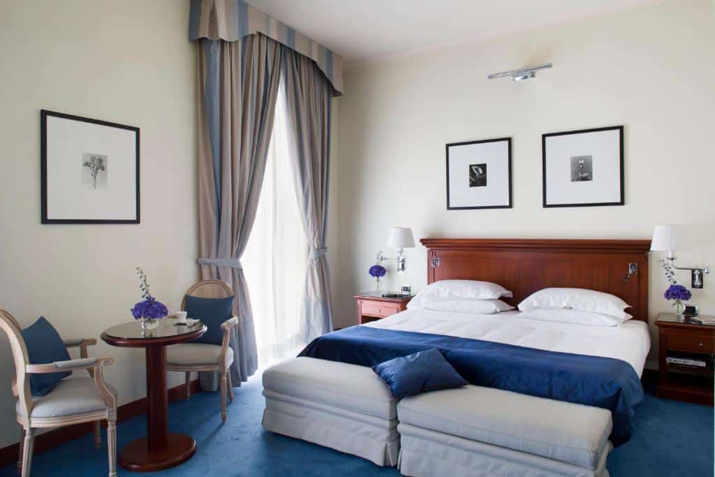 Quarto do Starhotels Terminus com uma cama de casal no meio do quarto, já em cada lado da cama tem uma mesinha com abajur e outros objetos, na direita há uma mesa com duas cadeiras e uma janela grande de vidro com cortinas.