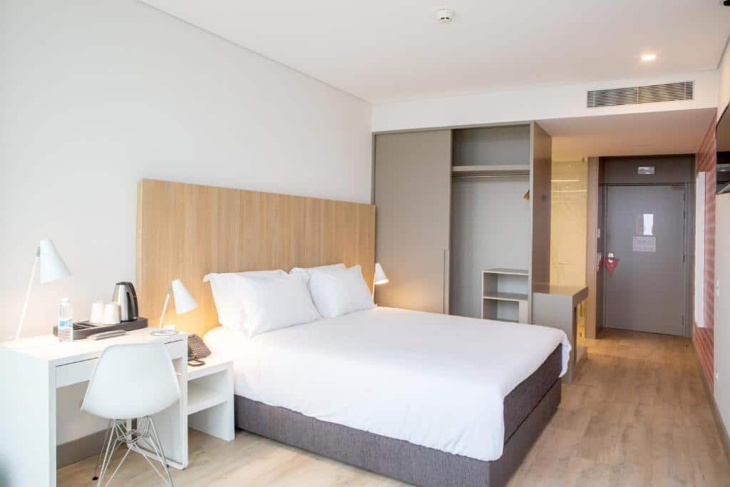 Quarto do Stay Hotel Porto Aeroporto com cama de sala do lado esquerdo da imagem com duas luminárias ao lado da cama e do lado esquerdo da cama uma mesa de trabalho com cadeira. Representa hotéis perto do aeroporto do Porto.