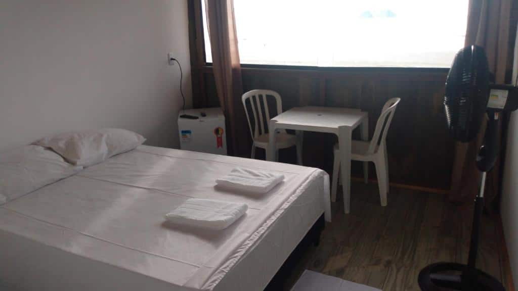 Quarto Suítes Pé na Areia. Uma cama de casal no lado esquerdo, atrás um frigobar e uma mesa de plástico com duas cadeiras. No lado direito um ventilador. Atrás da mesa, a janela do quarto com vista para a praia.