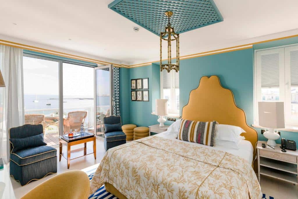 Quarto deluxe do  The Albatroz Hotel com uma cama de casal, ao lado esquerdo da cama há uma varanda de frente para o mar com duas cadeiras rústicas, também do lado esquerdo do quarto, há duas poltronas e uma mesinha de centro com comodidades para um café, para representar hotéis românticos em Lisboa