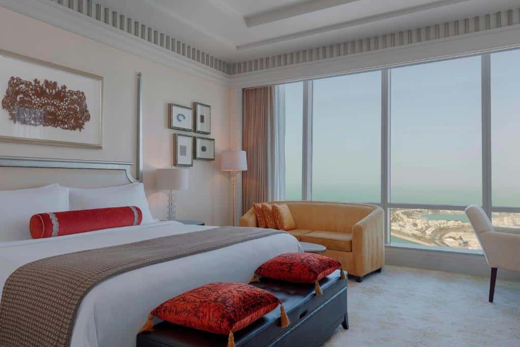Quarto do The St. Regis Abu Dhabi. Uma cama de casal no lado esquerdo, com uma almofada em cima, um sofá na frente com duas almofadas. No lado esquerdo da cama dois abajures e um sofá. No lado direito uma cadeira. No fundo, atrás do sofá, uma parede de vidro com vista para o mar e a cidade. Foto para ilustrar post sobre Hotéis em Abu Dhabi.