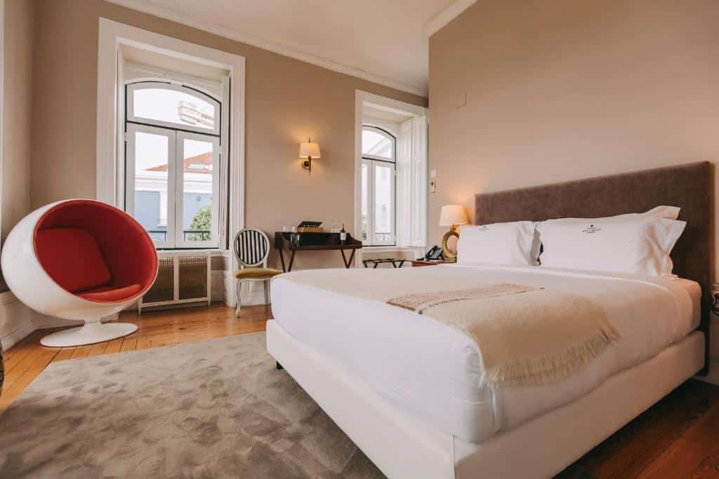 Quarto do Torel Palace Lisbon com uma cama de casal, duas janelas, entre elas há uma pequena mesa com um vinho e duas taças de vidro, há também um poltrona arredondada e um tapete