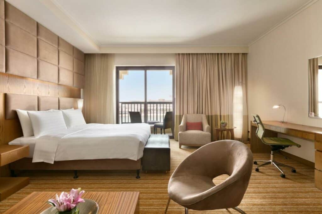 Quarto do Traders Hotel, Abu Dhabi. Uma cama de casal no lado esquerdo e um sofá na frente. De frente para a cama uma mesa de trabalho. Na frente da imagem uma mesinha de madeira e uma poltrona. No fundo uma poltrona com uma mesinha e a varanda do quarto, com duas cadeiras.