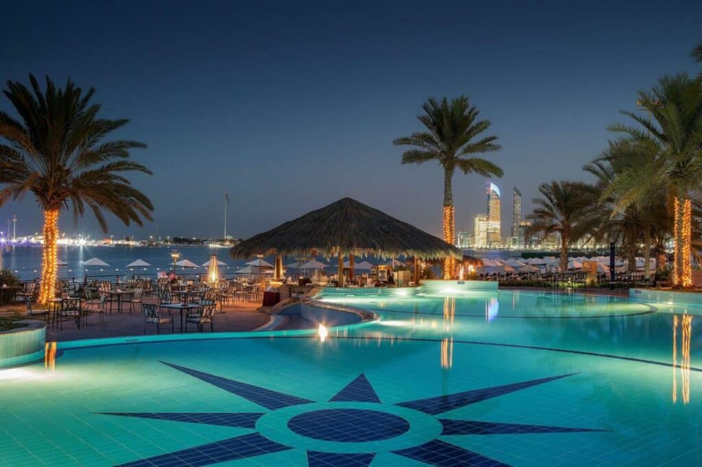 Piscina do Radisson Blu Hotel & Resort, Abu Dhabi Corniche. Uma piscina ao ar livre, do lado esquerdo cadeiras com cadeiras e guarda-sóis, Um quiosque beirando a piscina, palmeiras e no fundo, o mar.