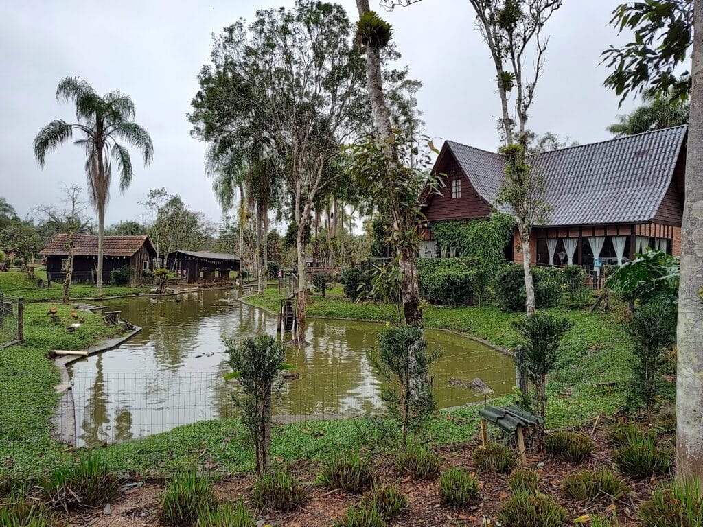 paisagem rústica do Rancho Alegre com um belo lago ao meio, algumas construções de casas de madeira ao redor, várias árvores e um dia cinzento.