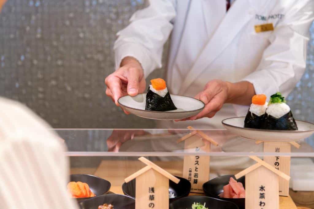 Foto tirada de um chef servindo sushi a um hóspede no Asakusa Tobu Hotel. Podemos ver parte da vitrine com vários pratos japoneses dentro.