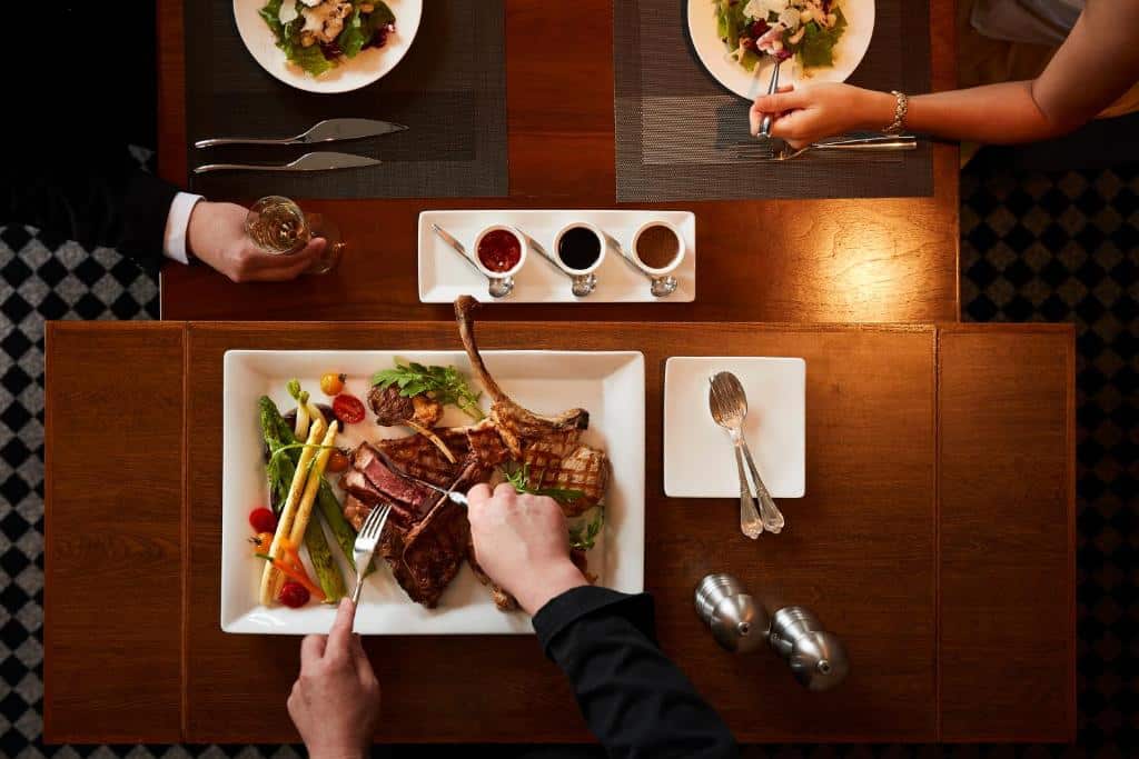 Vista de cima em uma mesa no hotel Grand NIkko Tokyo Daiba. É possível ver apenas os braços de três pessoas comendo em uma mesa, cada uma delas com um prato diferente. Podemos identificar saladas, carnes, vegetais e molhos. Um dos homens está segurando uma taça.