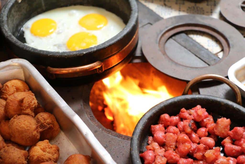 Imagem de um fogão à lenha na Pousada Nossa Senhora Aparecida, vemos alguns alimentos, como ovos, sendo preparados nas panelas.