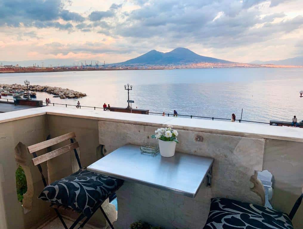 Parte de fora do Relais sul Mare Boutique Hotel com uma mesa e duas cadeiras de frente para o mar com algumas pessoas passeando, ilustrando post Hotéis em Nápoles.
