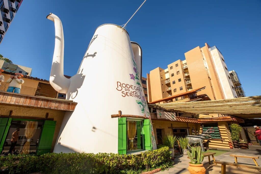Frente do restaurante Bodega do Sertão, onde há um bule enorme branco escrito "bodega do Sertão", além de vasos de flores e uma escultura de burro ao lado. Representa um dos restaurantes em Maceió