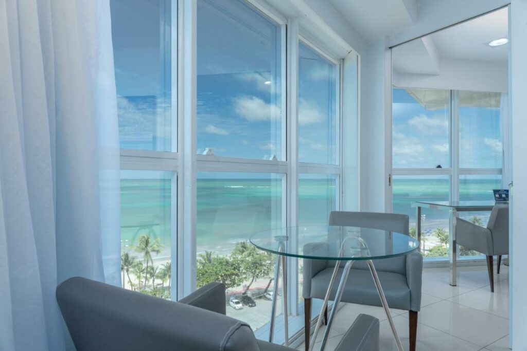 Área interna do hotel Brisa Praia, com mesa redonda de vidro com poltronas ao lado. e paredes de vidro ao lado e vista panorâmica do mar