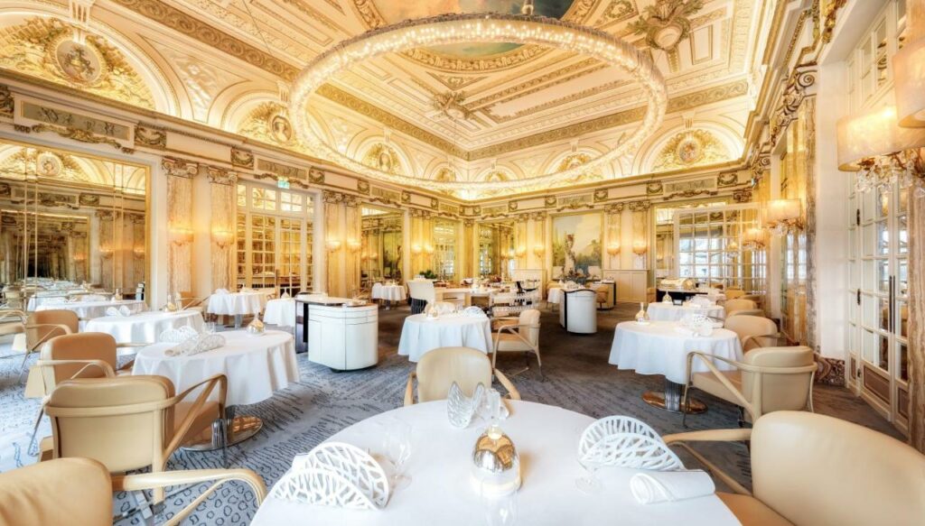 Restaurante do Hôtel de Paris Monte-Carlo. Várias mesas redondas com toalhas brancas e poltronas. O teto é dourado com iluminação amarela.