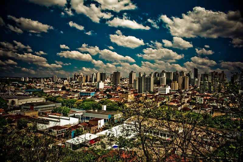 Paisagem da cidade de Ribeirão Preto, em São Paulo. Temos a visão de alguns galhos de árvores perto da câmera, casas, comércios e prédios no fundo. Está de dia e o céu possui várias nuvens.