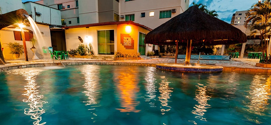 Parte do hotel Rio das Pedras Thermas Hotel que mostra a piscina com uma cascata e um quiosque no meio, e outras áreas da acomodação em volta, ilustrando post Hotéis em Caldas Novas.