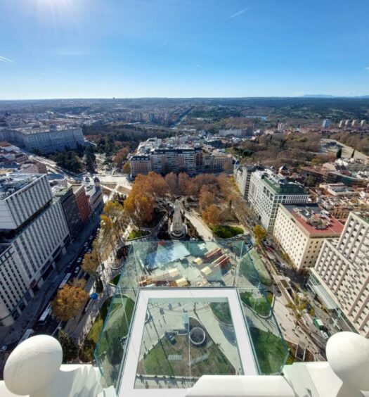 Vista de cima de Madri no Riu Plaza España, um dos melhores hotéis em Madri. A plataforma de vidro tem vista 360º para a cidade com seus prédios e praças arborizadas. O céu está azul na parte de cima da imagem, e o sol brilha no canto esquerdo. Logo abaixo do local fica a Plaza de España.