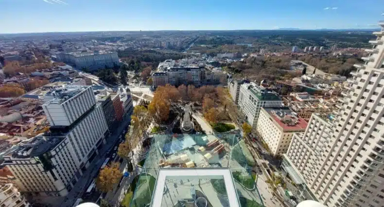 Vista de cima de Madri no Riu Plaza España, um dos melhores hotéis em Madri. A plataforma de vidro tem vista 360º para a cidade com seus prédios e praças arborizadas. O céu está azul na parte de cima da imagem, e o sol brilha no canto esquerdo. Logo abaixo do local fica a Plaza de España.