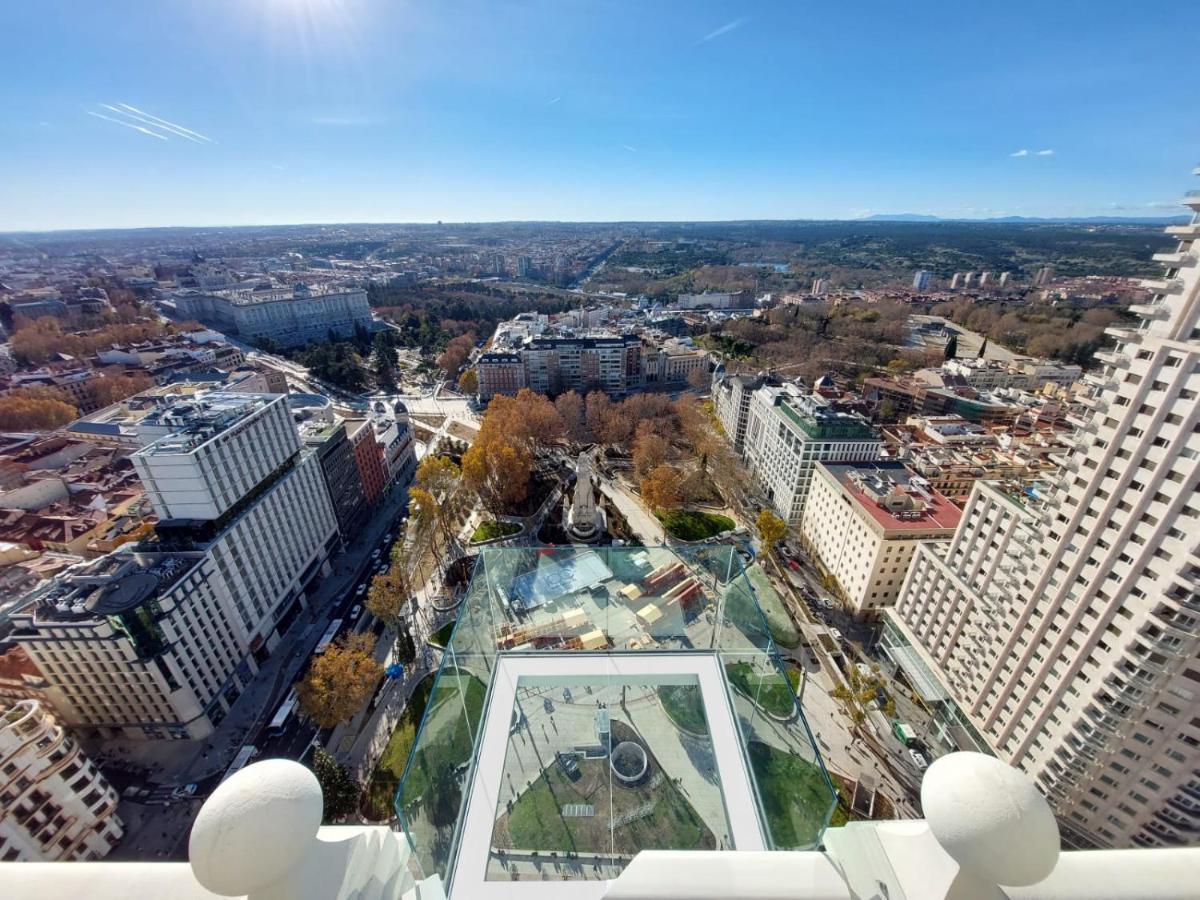 Vista de cima de Madri no Riu Plaza España, um dos melhores hotéis em Madri. A plataforma de vidro tem vista 360º para a cidade com seus prédios e praças arborizadas. O céu está azul na parte de cima da imagem, e o sol brilha no canto esquerdo. Logo abaixo do local fica a Plaza de España.