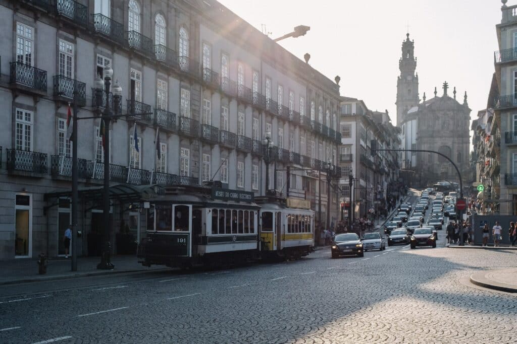 Rua em Porto durante o dia com carros circulando do lado direito e do lado esquerdo um bondinho. Representa aluguel de carros no aeroporto do Porto.