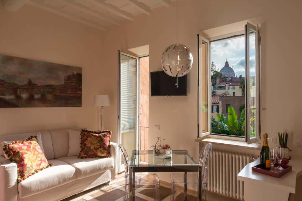 sala do La Porta Rossa di Borgo - Vatican Luxury Suite, com janela que dá vista para a Basílica de São Pedro, varanda, sofá, mesinha no centro de vidro com cadeiras e tapete, há uma luminária de teto em formato de globo de festa