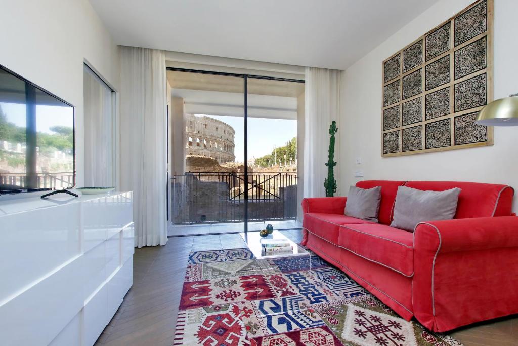 sala do Luxury Domus Apartment 1 com tapete colorido e mesinha de centro transparente, há um sofá vermelho e tv à frente com bancada branca e, do outro lado, uma janela grande com cortinas que dá vista direta para o Coliseu