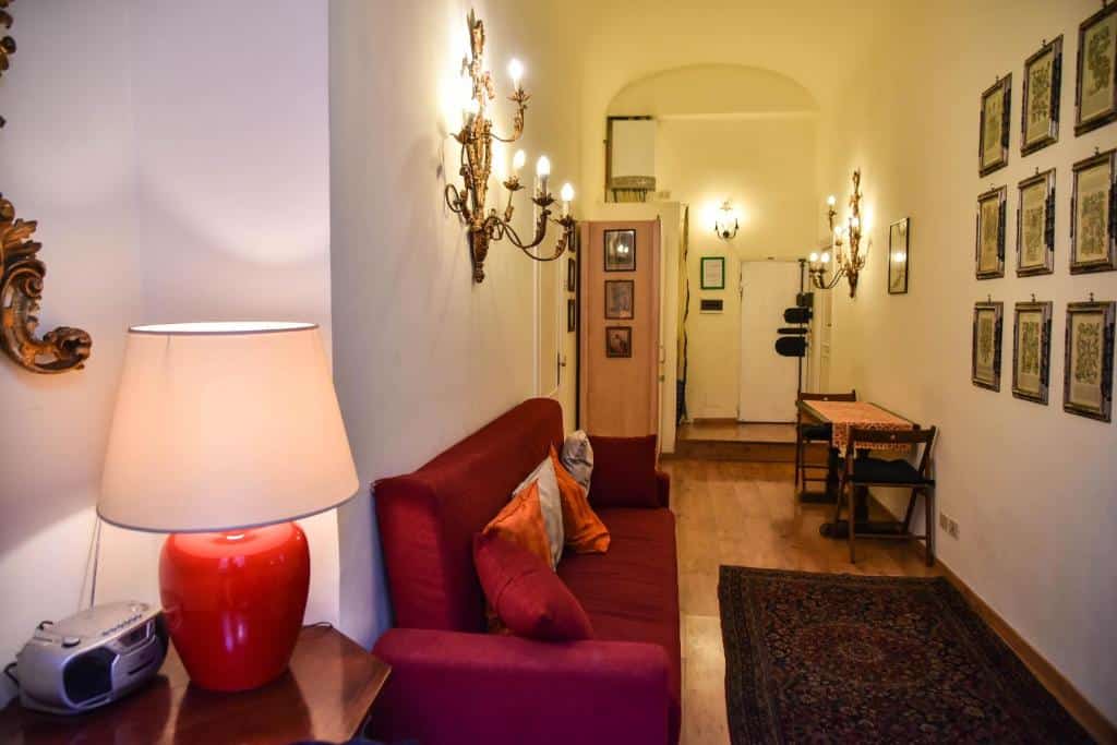 sala do Stylish apartment in central Rome "Centro Storico", com sofá vermelho com almofadas, mesinha com luminária e rádio, com mesa e cadeiras e outros móveis de madeira, há candelabros nas paredes e tapete no chão em um ambiente comprido