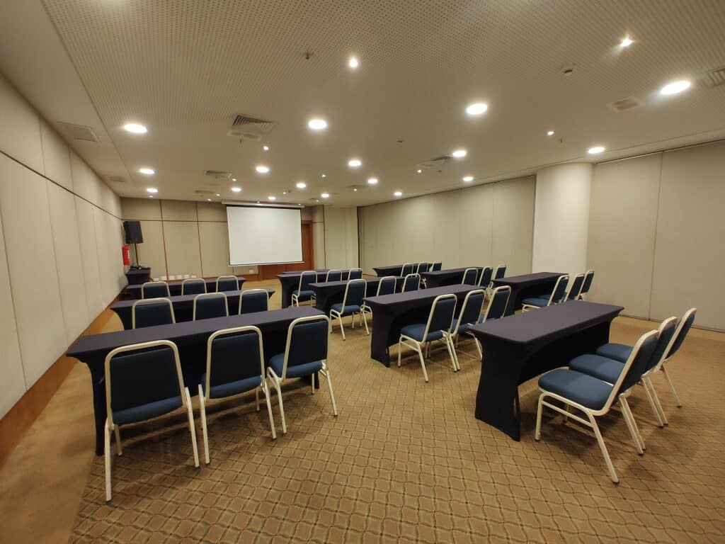 Sala de conferência com várias mesas estreitas, encapadas com um tecido azul escuro, e com três cadeiras estofadas, também azuis, em cada mesa. No fundo da sala é possível ver uma tela branca de projetor puxada para baixo.