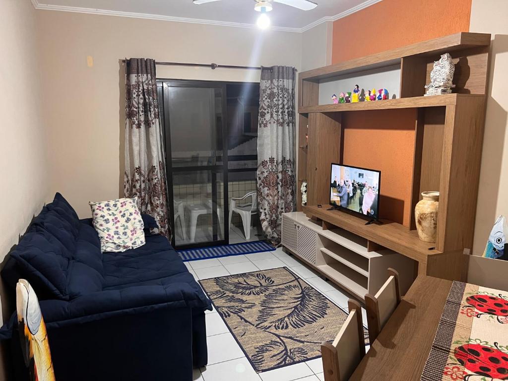 Sala de apartamento com sofá no lado esquerdo, mesa, cadeiras e móvel com televisão no lado direito, tapete no meio da sala, porta de vidro com cortina que vai para a varanda.