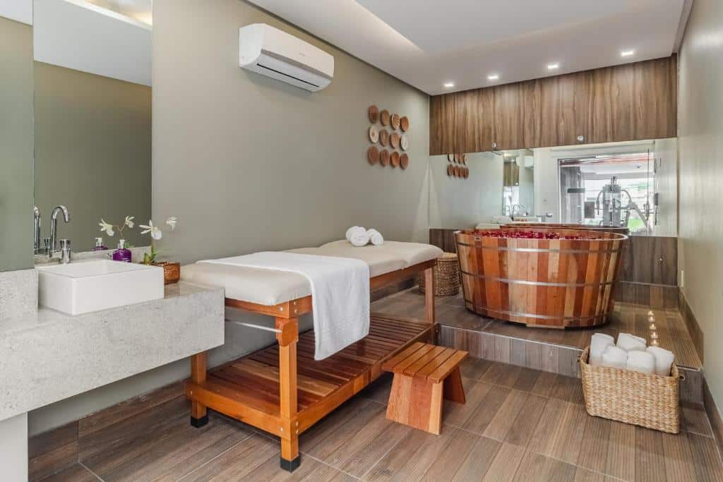Área de spa do Citi Hotel Premium Caruaru. No lado esquerdo há uma pia, um espelho e uma maca. Já ao fundo há uma banheira de madeira com pétalas de rosas e um espelho atrás da banheira.