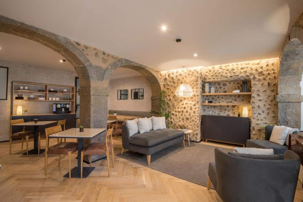 Área compartilhada do Urbano FLH Hotels Lisboa com sofás, poltronas e uma televisão, o ambiente é dividido por paredes de concreto, há também mesinhas quadradas e cadeiras estofadas, para representar hotéis no Chiado em Lisboa
