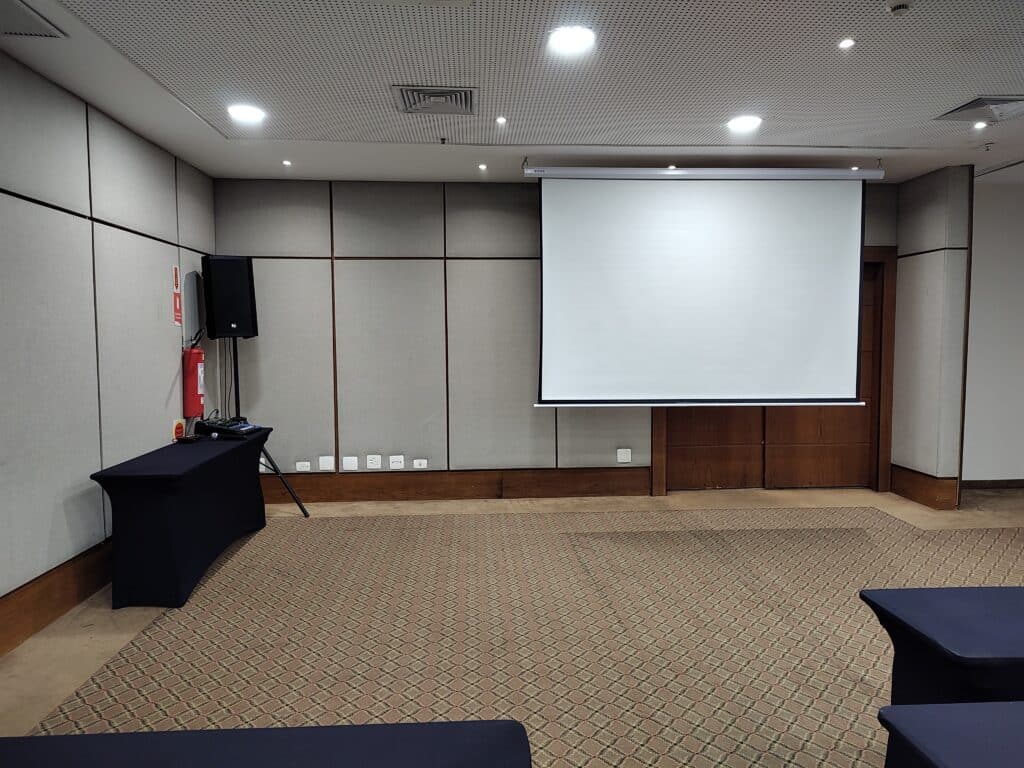 sala de reunião do Bourbon Joinville Convention Hotel mostrando uma tela de projeção em branco na parede do fundo. No canto esquerdo da sala há uma mesa estreita com uma pequena mesa de som e um microfone em cima