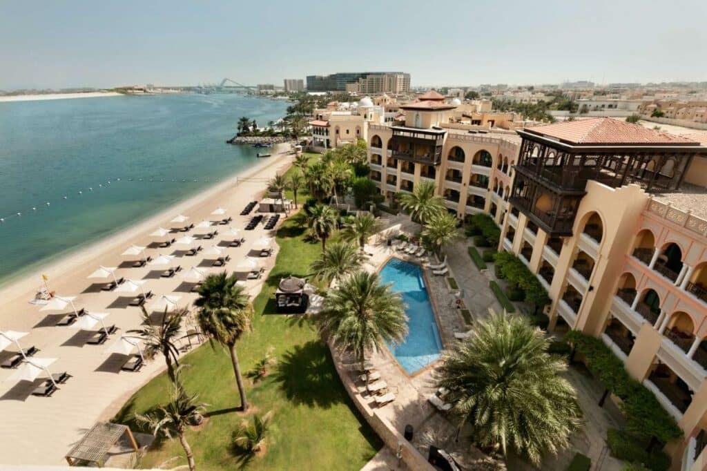 Vista de cima do Shangri-La Qaryat Al Beri, Abu Dhabi. Do lado direito o hotel, árvores e uma piscina com cadeiras de tomar sol ao redor. No meio, um jardim com árvores. No lado esquerdo a praia, cadeiras de tomar sol com guarda-sóis e o mar.