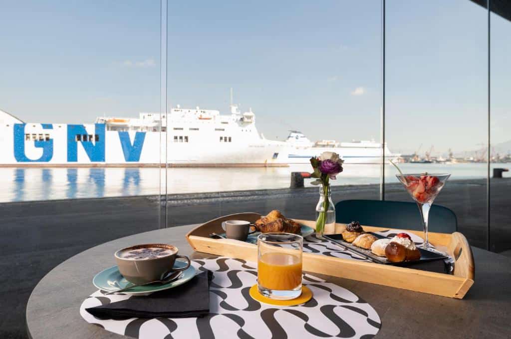 Mesa redonda do Smart Hotel Napoli com comidas em cima e com uma vista da parede de vidro para o navio branco parado no mar, ilustrando post Hotéis em Nápoles.