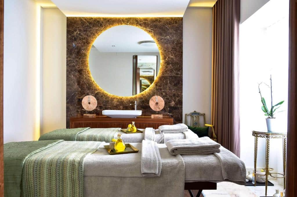 Área de spa do Tivoli Palácio de Seteais Sintra Hotel com duas macas com toalhas e um bandeja com óleos essenciais, uma ampla janela com cortinas e um espelho redondo na parede ao fundo, para representar hotéis românticos em Lisboa