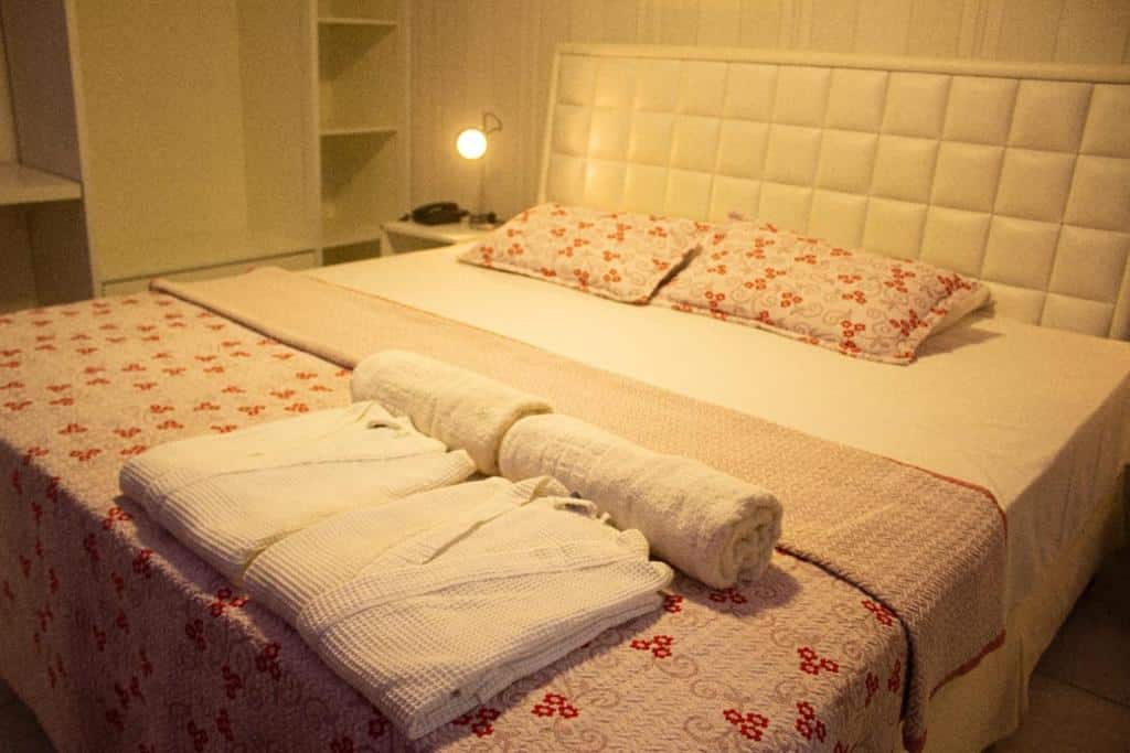 Quarto do Caruaru Pallace Hotel. A cama está centralizada, tem duas toalhas e dois roupões em cima, há um pequeno móvel no lado esquerdo da cama com um abajur ligado e ainda no lado esquerdo da cama há um armário. Imagem para ilustrar o post hotéis em Caruaru.
