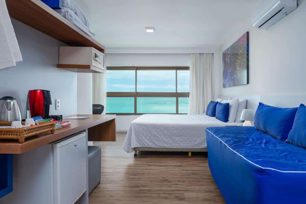 Suíte com vista do mar do hotel Ponta Verde Maceió, de 23 m², com um sofá azul e à esquerda um balcão com amenidades para chá e café, um frigobar em baixo e um cofre suspenso. Ao fundo, tem uma cama de casal e uma janela panorâmica com vista do mar