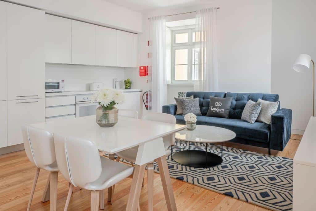 Sala de estar junto de uma cozinha no Tandem Palacio Alfama Suites, há também um sofá com almofadas e uma mesa para refeições branca com quatro cadeiras