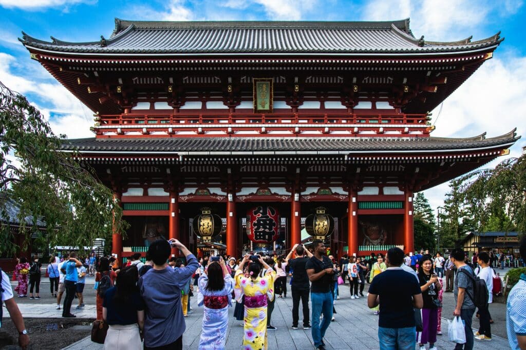 Pessoas visitando o Templo Senso-ji, em Asakusa, Tóquio. O templo está centralizado na foto, e várias pessoas tiram foto ao seu redor. Algumas mulheres usam roupas tradicionais japonesas.