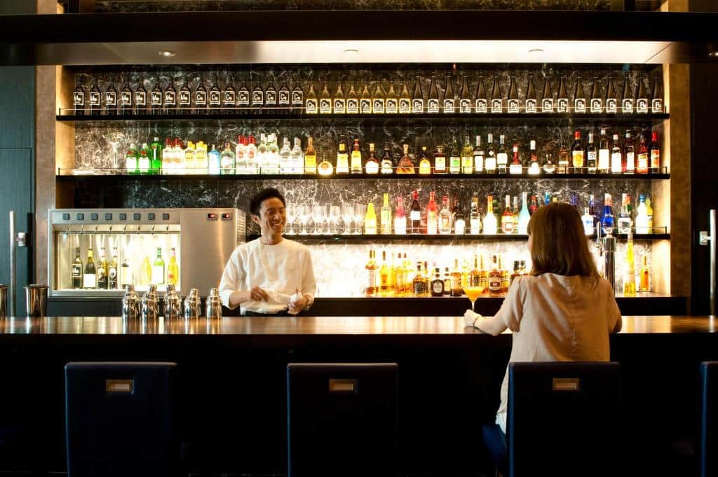 Foto do bar do The Gate Hotel Asakusa.O barman conversa com uma cliente, que segura uma taça na mão e está sentada no balcão. Há várias prateleiras iluminadas com bebidas destiladas e taças atrás do bar.